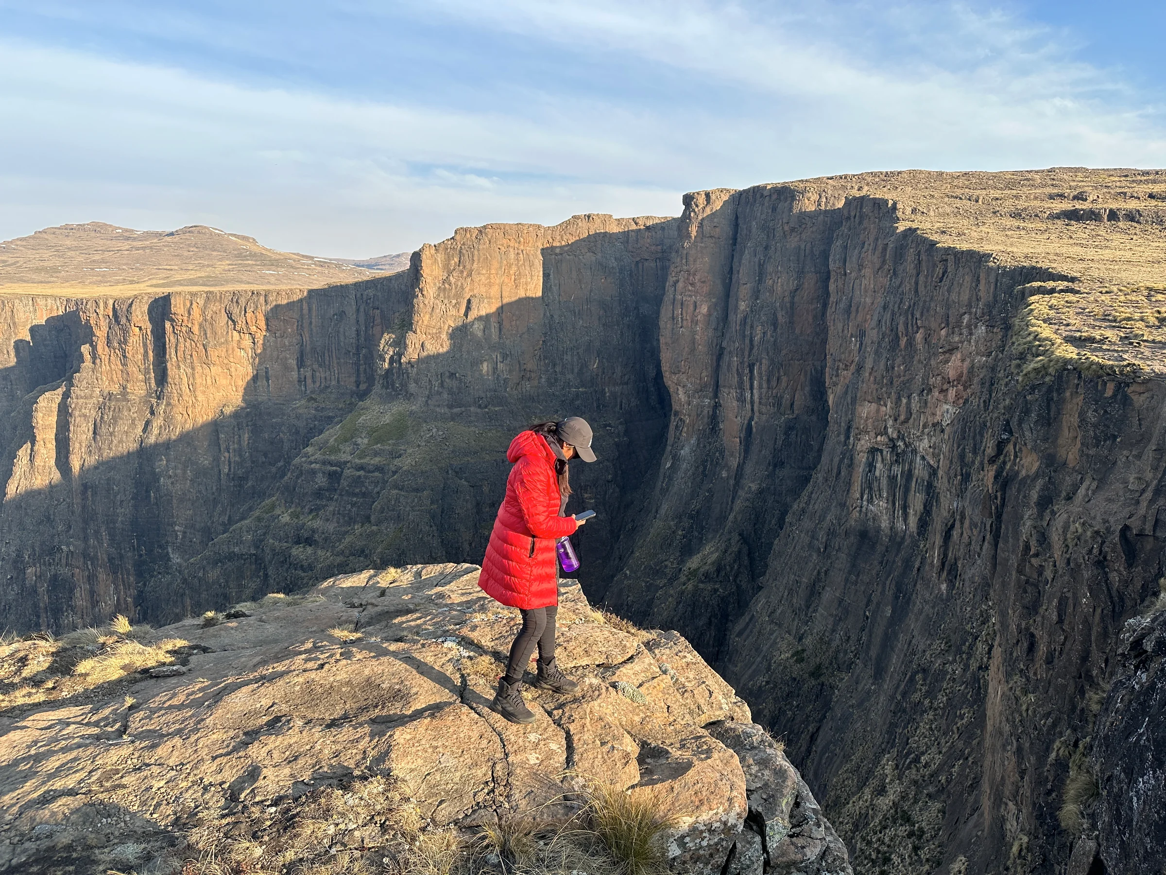 A hiker in a red coat on a rocky ledge above sunlit cliffs