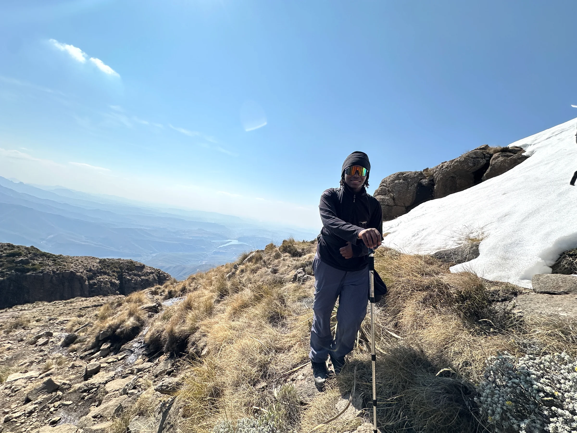 A hiker resting on a trekking pole beside a patch of mountain snow