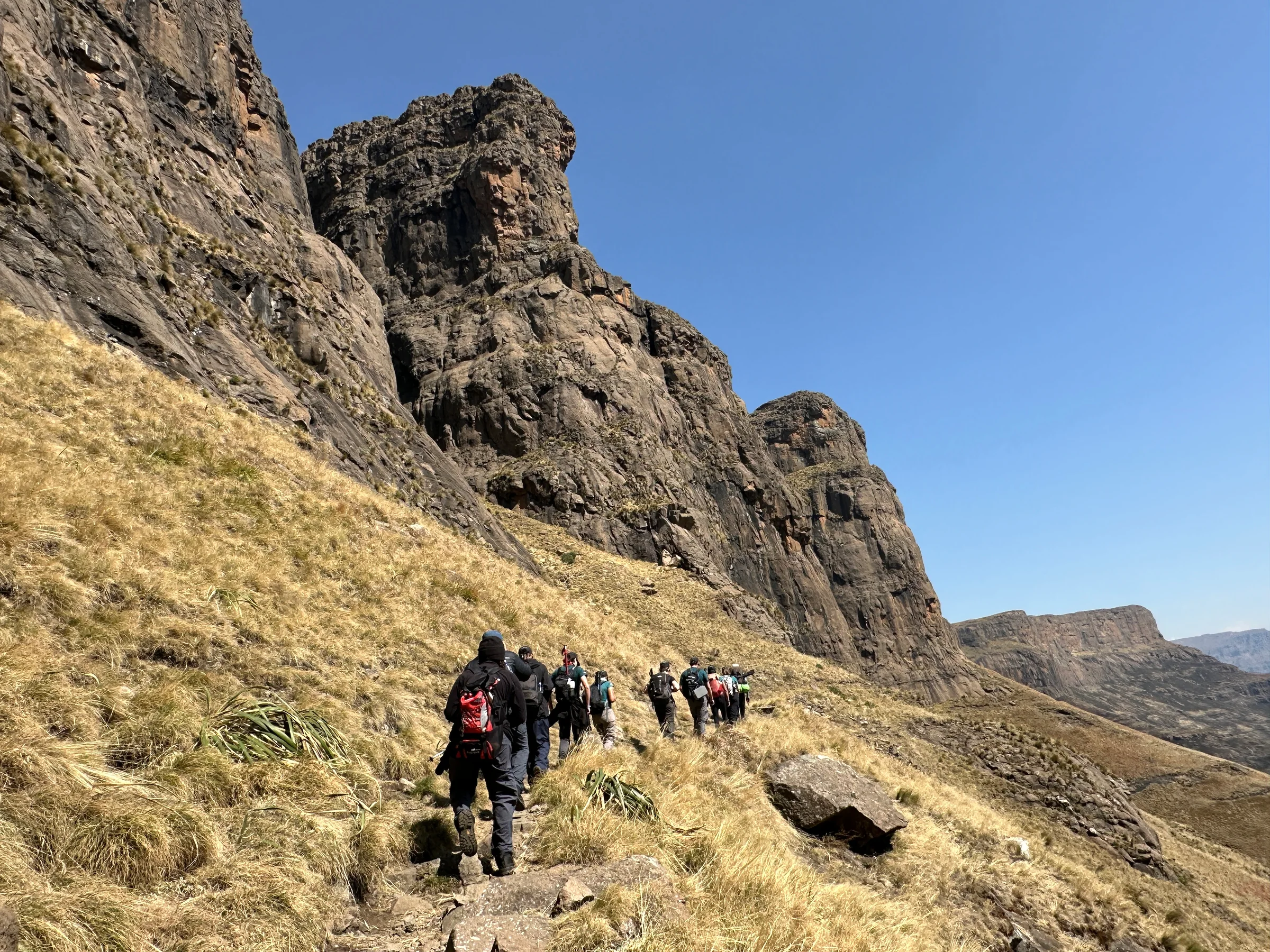 A group following a grassy mountain path beneath towering cliffs