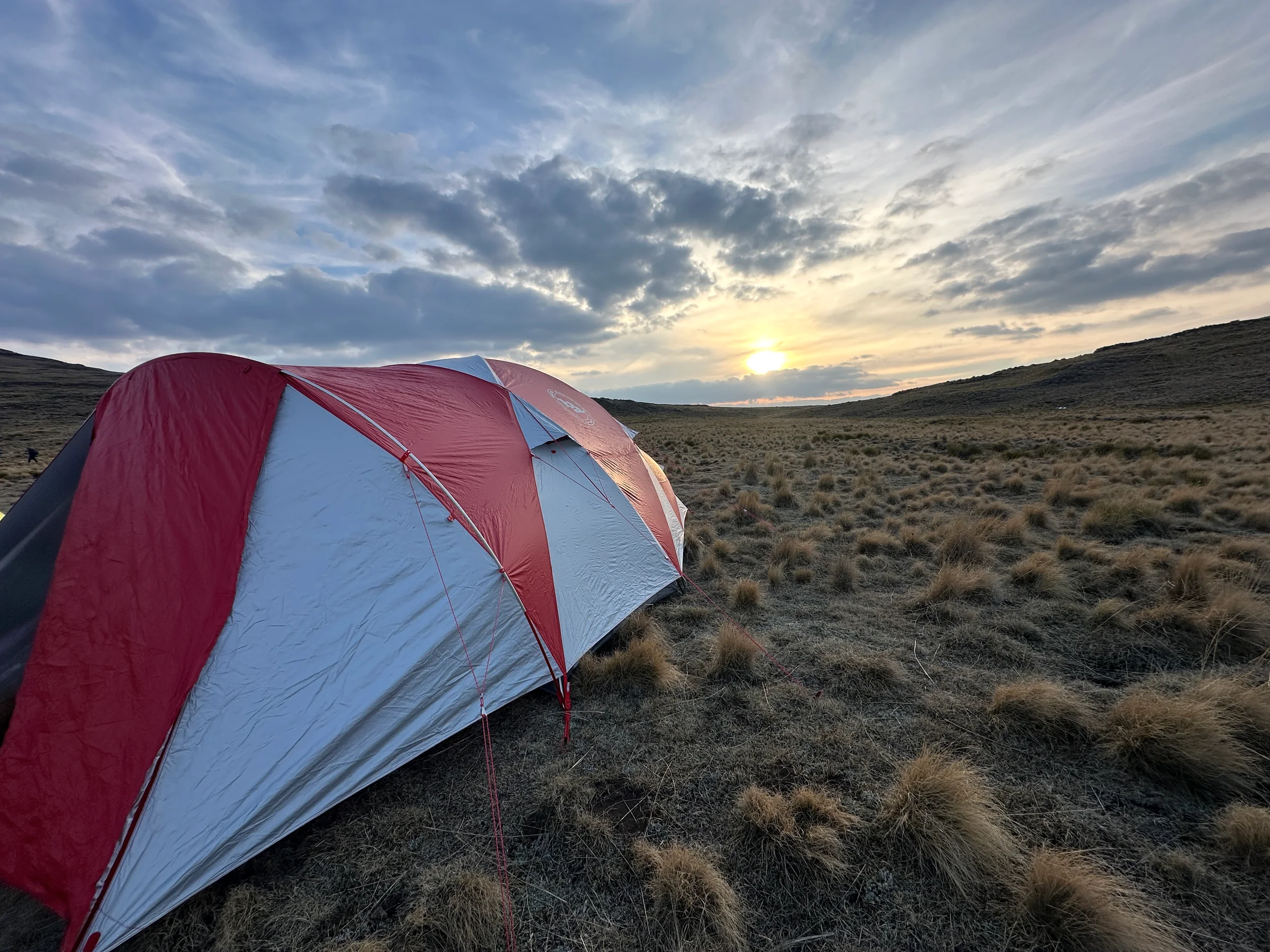 A red and grey tent on mountain grassland beneath sunset clouds